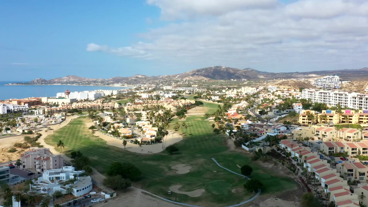Aerial view of coastal homes, golf greens, ocean, and hills under scattered clouds.