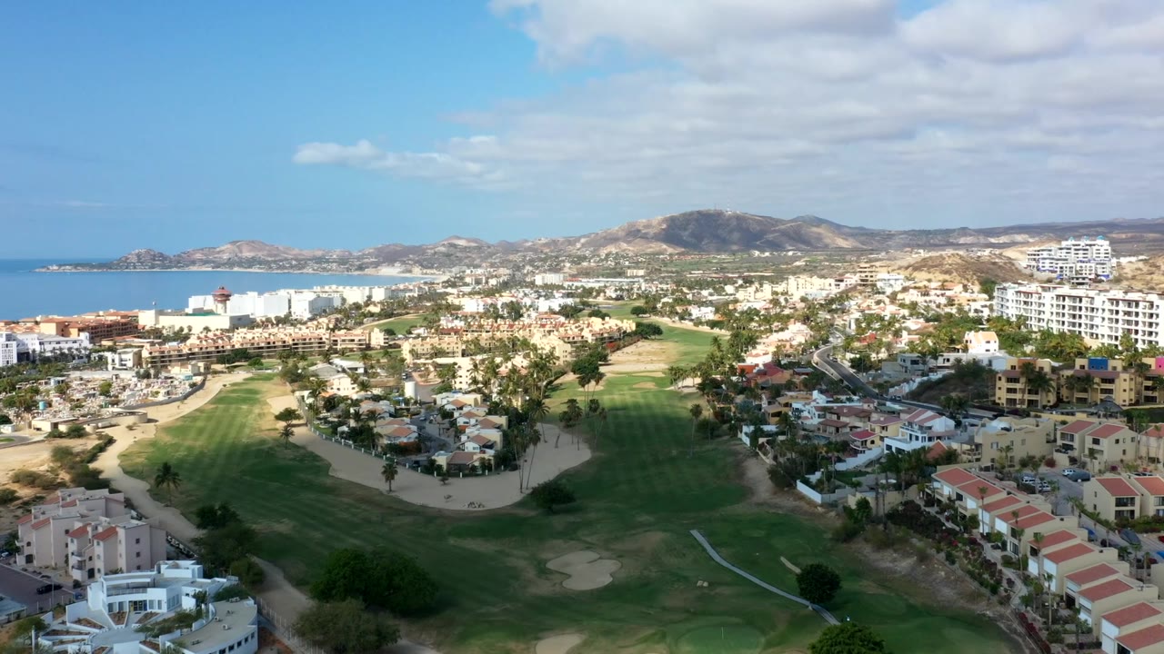Aerial panorama of ocean, resort-style buildings, golf course, roads, and hillside terrain.