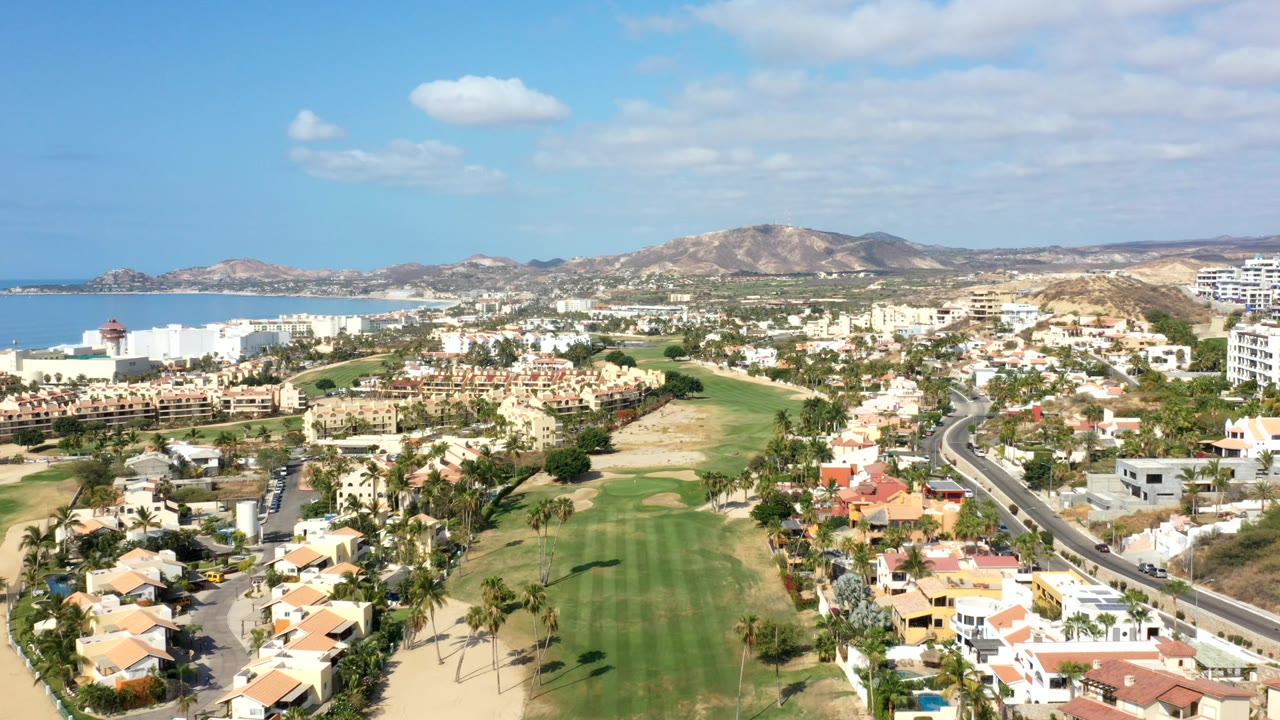 Opening aerial frame over Los Cabos, Mexico, showing green fairways, palm trees, residential buildings, blue water, dry hills, scattered clouds, and no visible people.