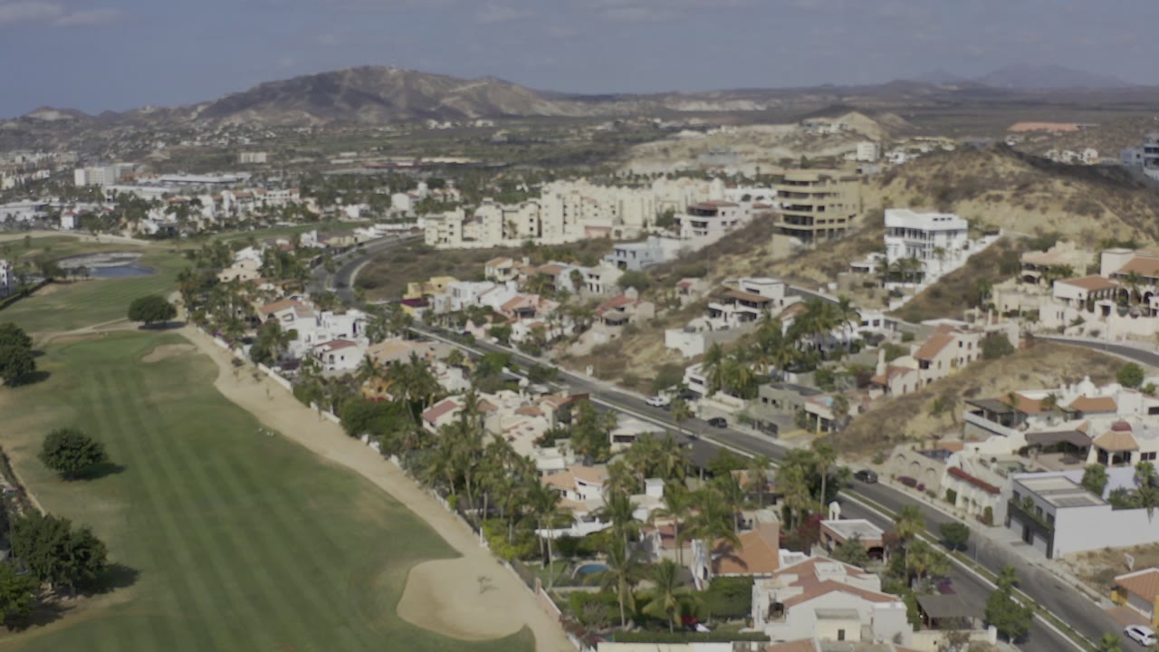 Opening Los Cabos aerial frame showing a narrow green fairway, sand trap, palm-lined homes, hillside buildings, distant mountains, and no visible people.
