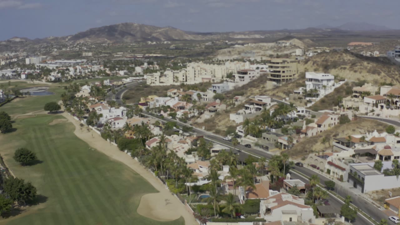 Los Cabos aerial frame with the long fairway on the left, a road through the middle, clustered homes below the hills, and no visible people.