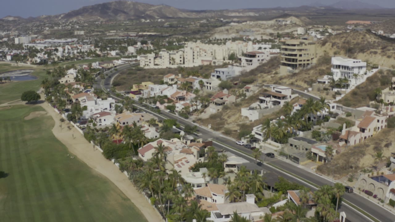 Start of the Los Cabos aerial view showing a green field at left, a curving residential road, pale homes, dry hillside lots, distant buildings, and no visible people.