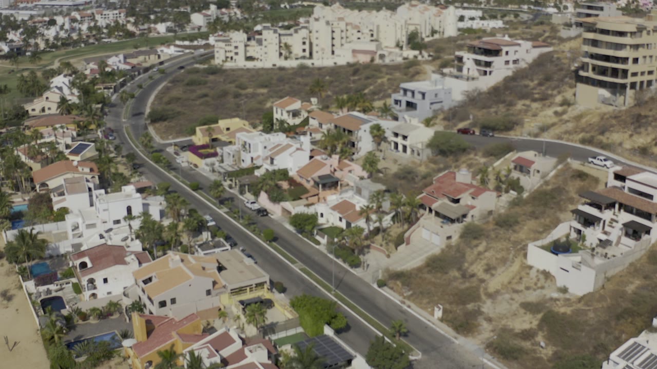 Aerial view over Los Cabos homes with a curving road at left, dry scrub lots at center, low-rise buildings in the background, and no visible people.