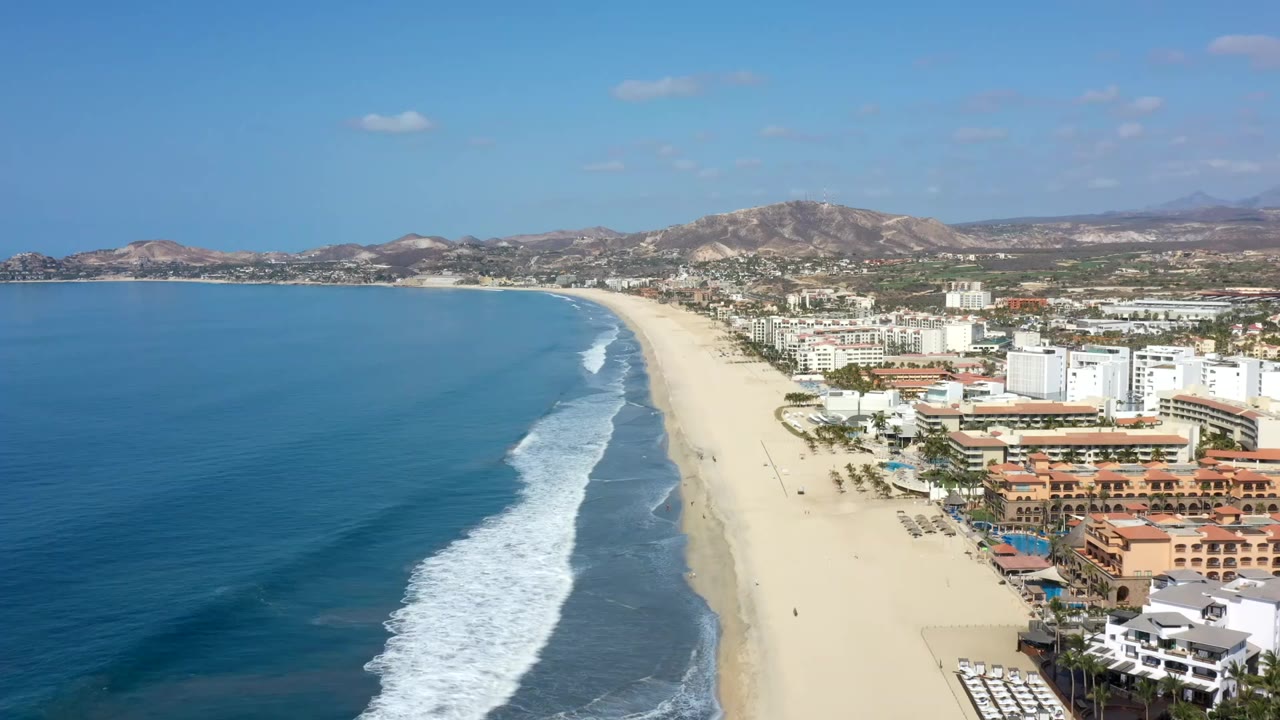 Aerial view of blue ocean, white surf, long beach, resort buildings, and inland hills.