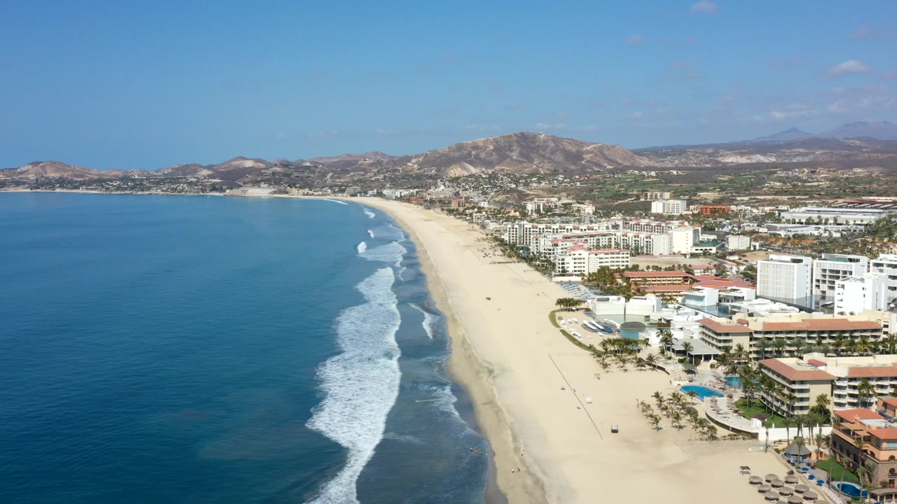 Aerial view over a Los Cabos beach with deep blue ocean on the left, white surf near shore, beachfront condos on the right, dry hills beyond, and no visible people.