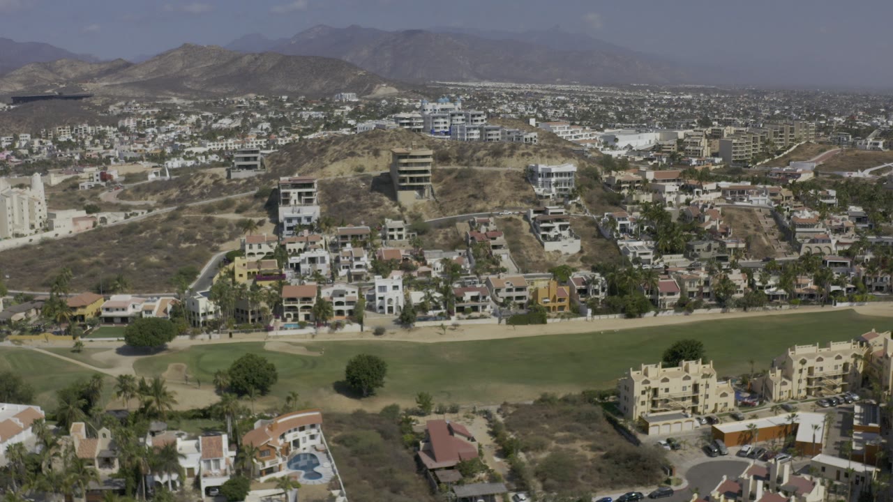 Opening aerial frame in Los Cabos, Mexico, looking across green parkland toward dry hills, residential buildings, and a hazy mountain horizon with no visible people.