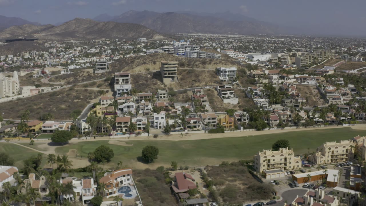 Aerial frame in Los Cabos, Mexico, with a broad green lawn at the bottom, clustered homes and apartment blocks across dry slopes, and no visible people.