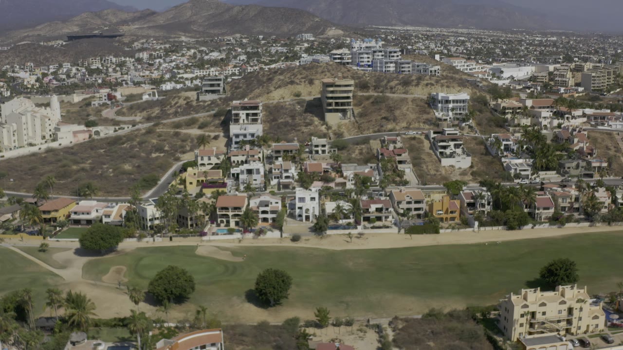 Los Cabos aerial frame with villas and apartment-style buildings above a broad green field, dry hillside roads, hazy mountains, and no visible people.