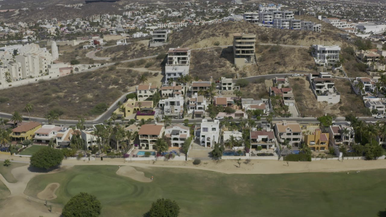 Aerial frame over Los Cabos hillside homes, apartment buildings, roads, green golf course turf, and sand traps, with no visible people.