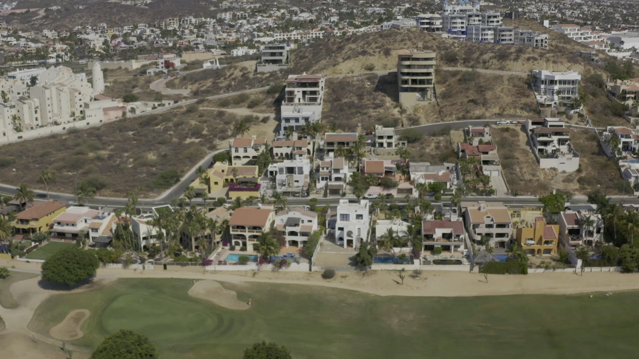 Mid-clip aerial frame over Los Cabos residences, with golf course turf and bunkers below, clustered homes in the middle, hillside apartments behind, and no visible people.