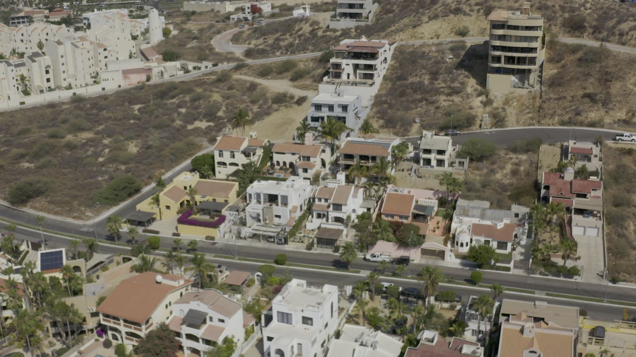 Aerial view over a Los Cabos residential hillside with white homes, tan roofs, a paved road, dry open land, parked vehicles, and no visible people.