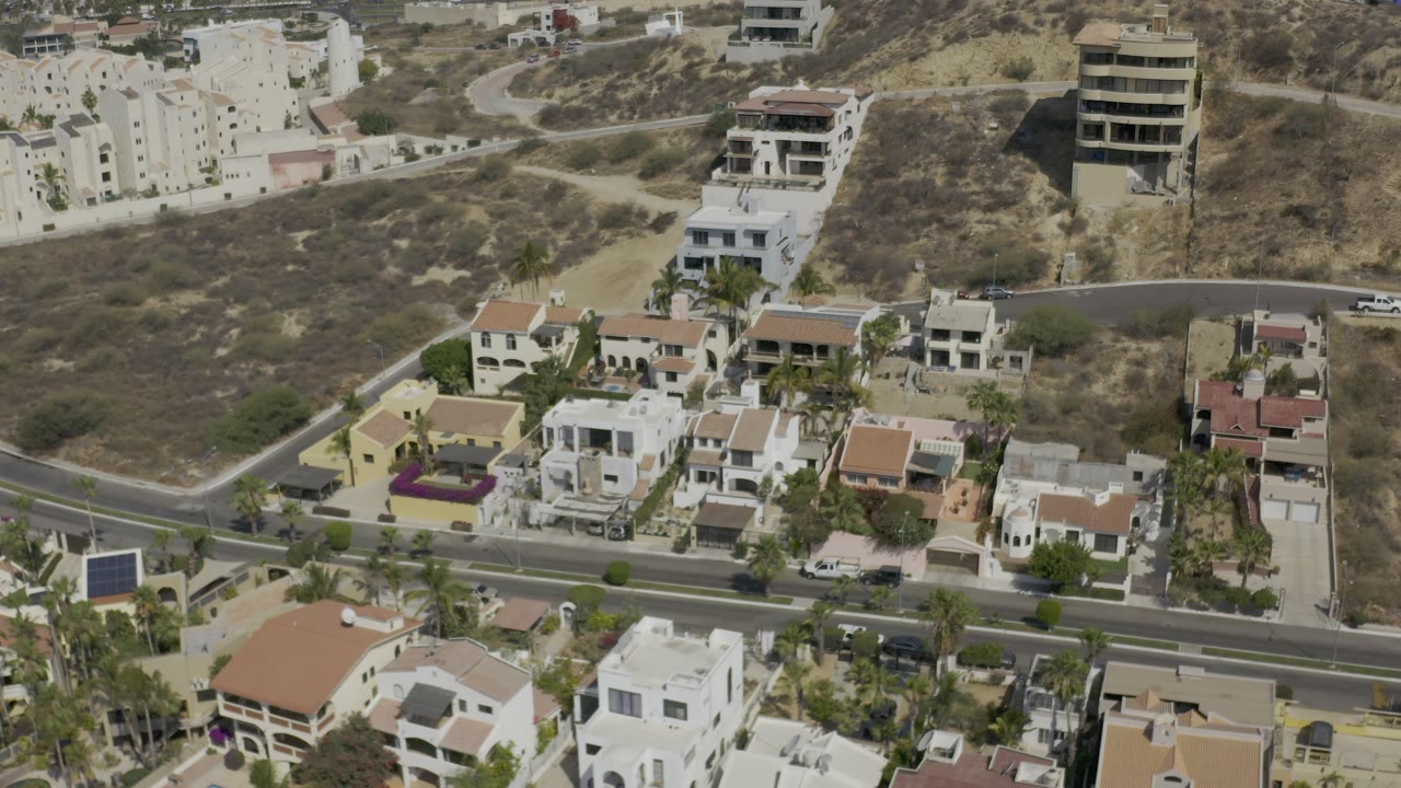 Elevated Los Cabos view with clustered homes in the foreground, a dark paved road across the frame, dry hillside lots behind, and no visible people.