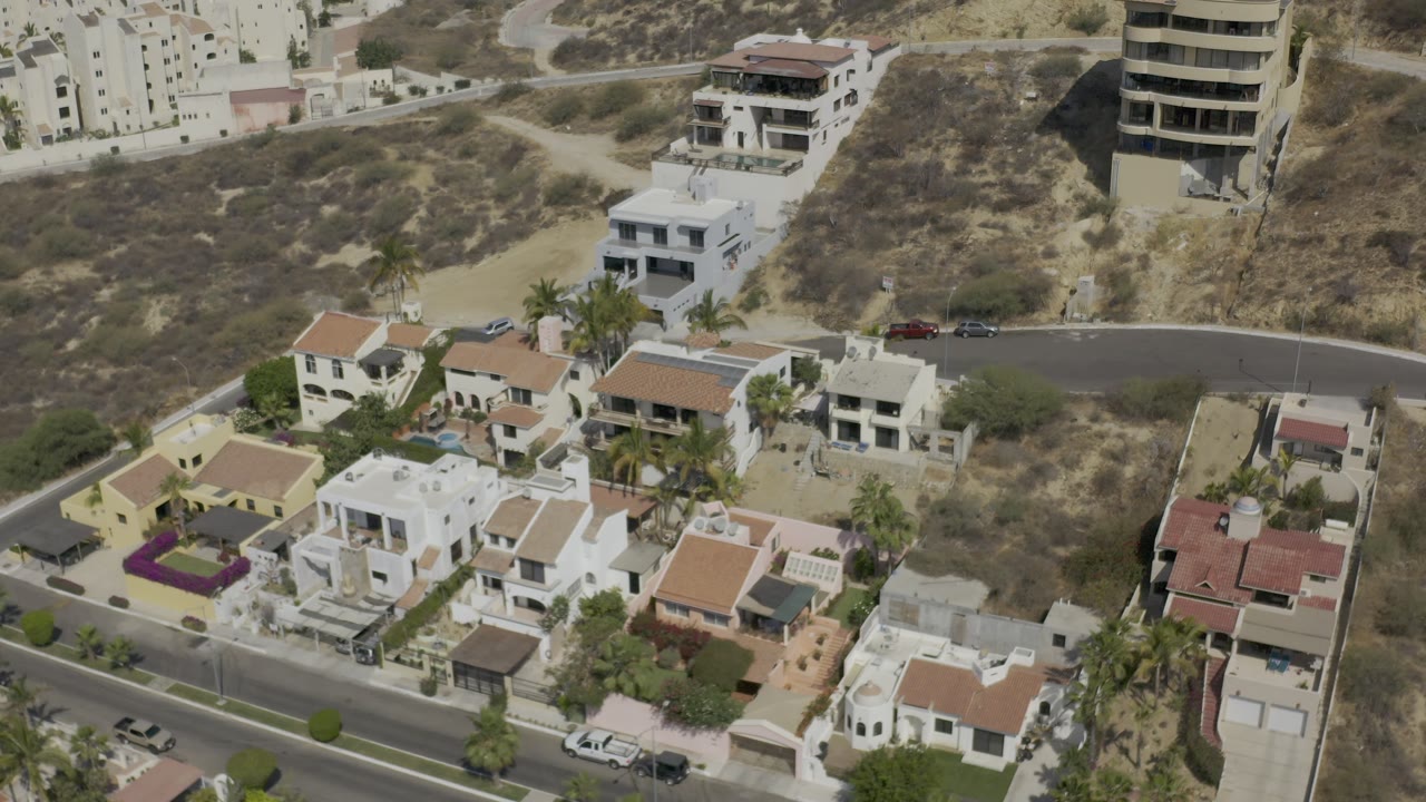 Los Cabos aerial view showing a curved road above clustered houses, dry hillside scrub in the background, palm trees among the homes, and no visible people.