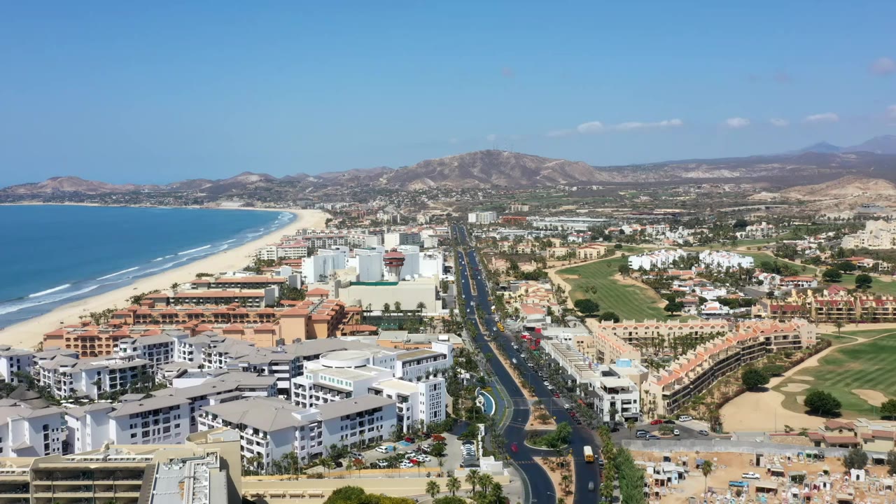 Wide aerial frame over Los Cabos, Mexico, with blue ocean and sandy beach at left, a divided road through the center, hotels and low buildings below, dry hills beyond, and no visible people.