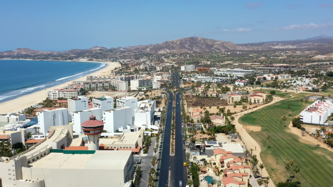 Aerial view of a coastal city with beach, road, buildings, golf course, and hills.