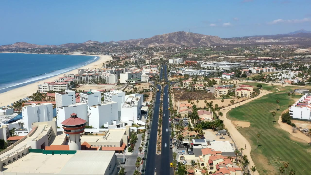 Drone view of beachside buildings, road, golf course, and arid hills.