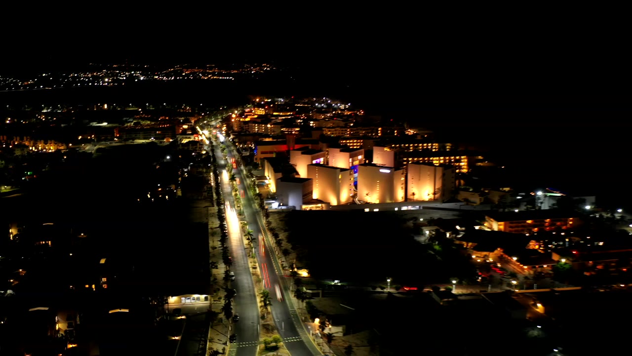 Aerial night frame over Los Cabos, Mexico, with a bright diagonal roadway, warm-lit tall buildings to the right, distant city lights, and no visible people.