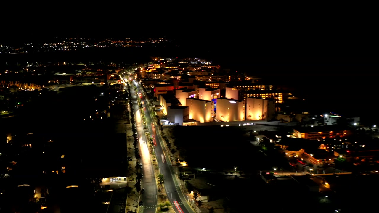 Los Cabos night aerial with the road centered more tightly, white headlights and red taillight streaks, bright building facades on the right, and no visible people.
