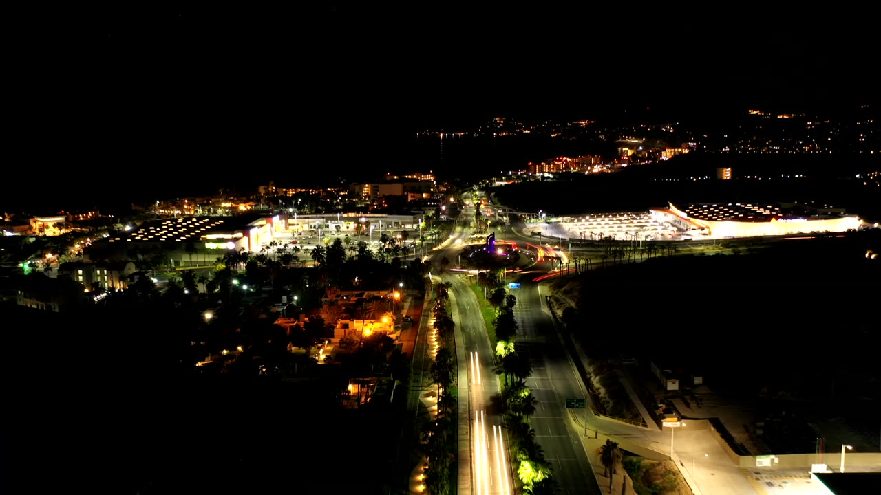Opening frame of Los Cabos, Mexico at night from an elevated view, with bright buildings, roads, car light streaks, and no visible people.