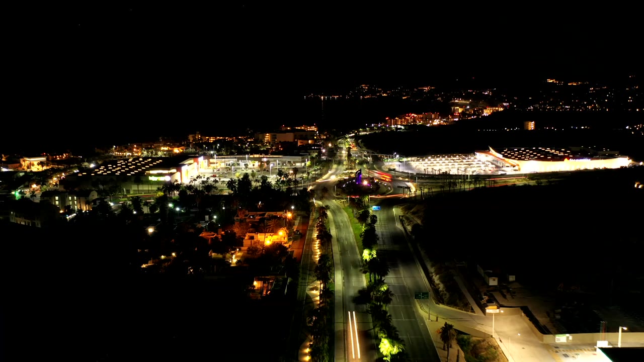Los Cabos night aerial with a bright building complex to the right, glowing road lanes in the center, distant lights, and no visible people.