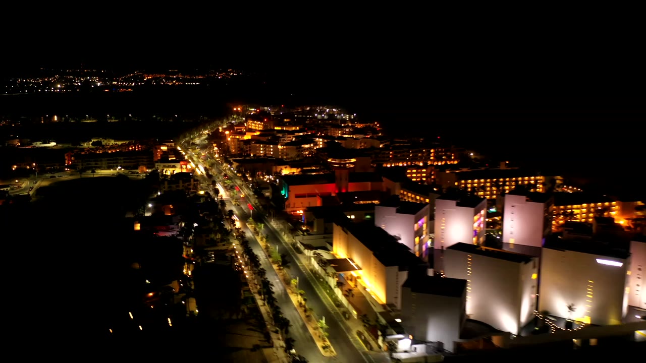 Elevated nighttime view in Los Cabos, Mexico, with cars on a diagonal boulevard and warm-lit building facades, no visible people.