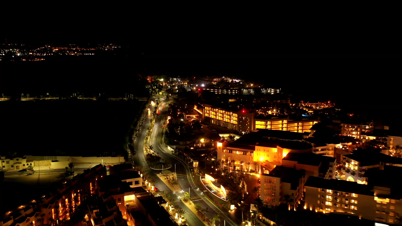Night drone frame over Los Cabos, Mexico, with a lit roadway curving beside dark water, warm building lights, distant shoreline lights, and no visible people.