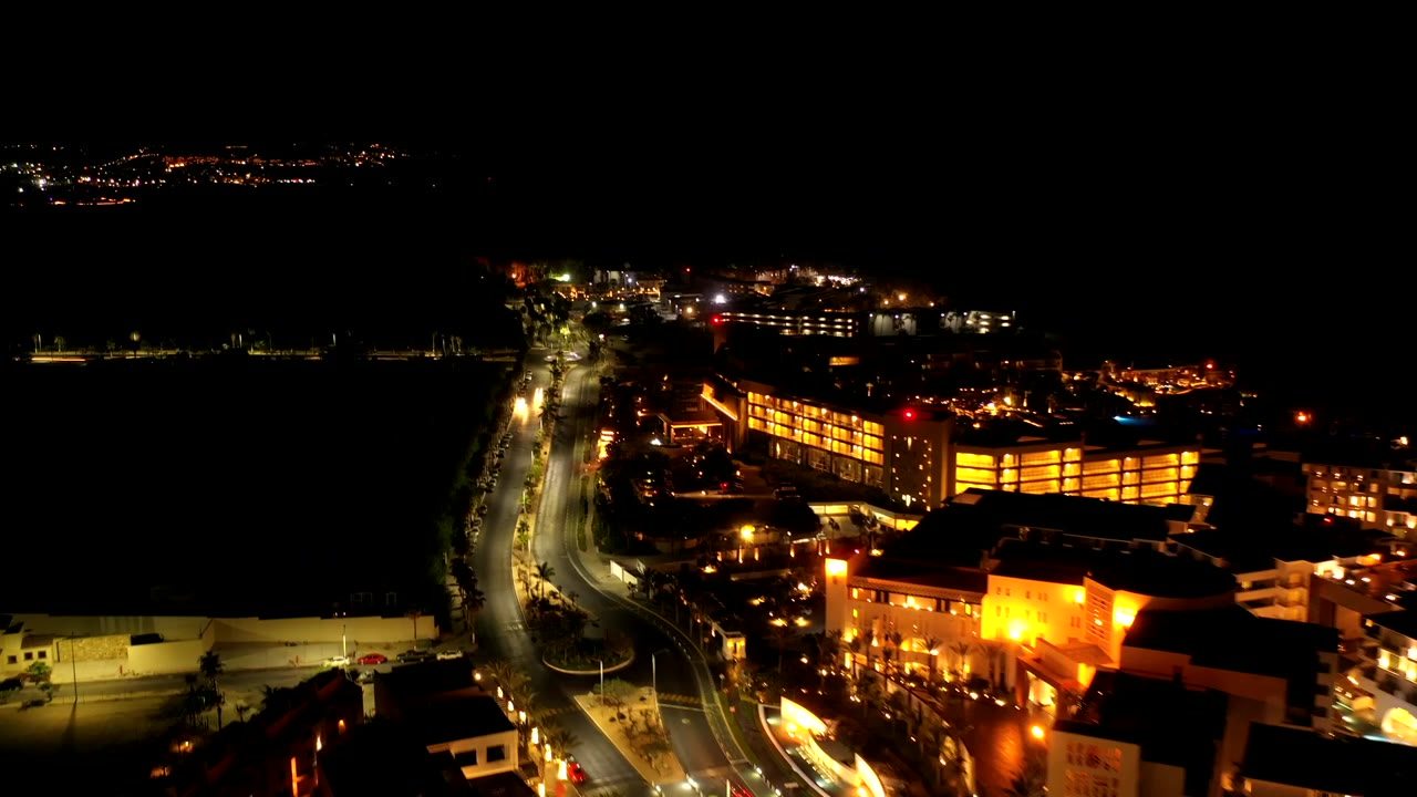 Drone view of a waterfront street with bright buildings at night.