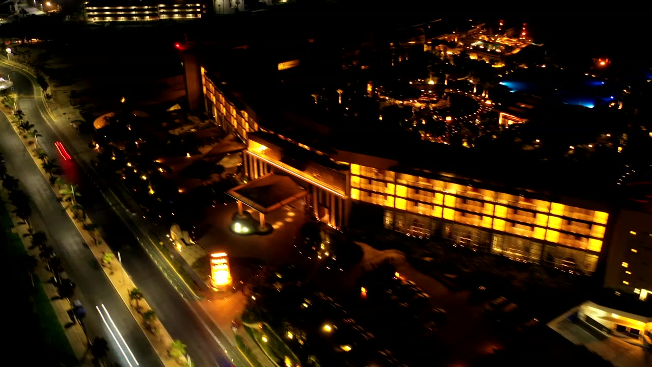 Elevated Los Cabos night view with the lit window grid stretching across frame, the street along the left edge, and no visible people.
