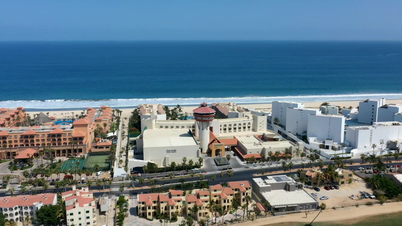 Wide aerial frame of Los Cabos, Mexico, showing terracotta and white coastal buildings, a sandy beach, blue ocean bands, and no visible people.