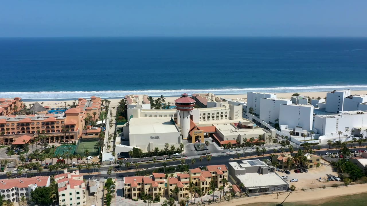 Aerial view in Los Cabos, Mexico, with the coastline stretching behind white resort-scale buildings, terracotta condo blocks, palm trees, and no visible people.