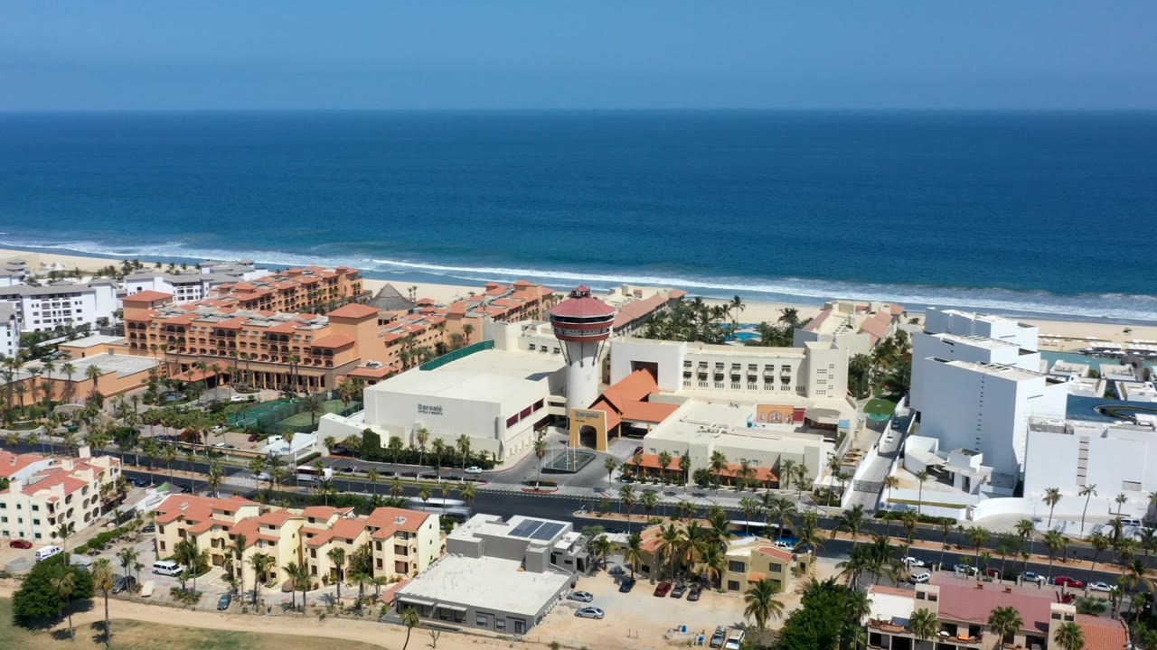 Aerial frame of Los Cabos with blue ocean and surf beyond beachfront buildings, a palm-lined road, red-roofed homes, and no visible people.