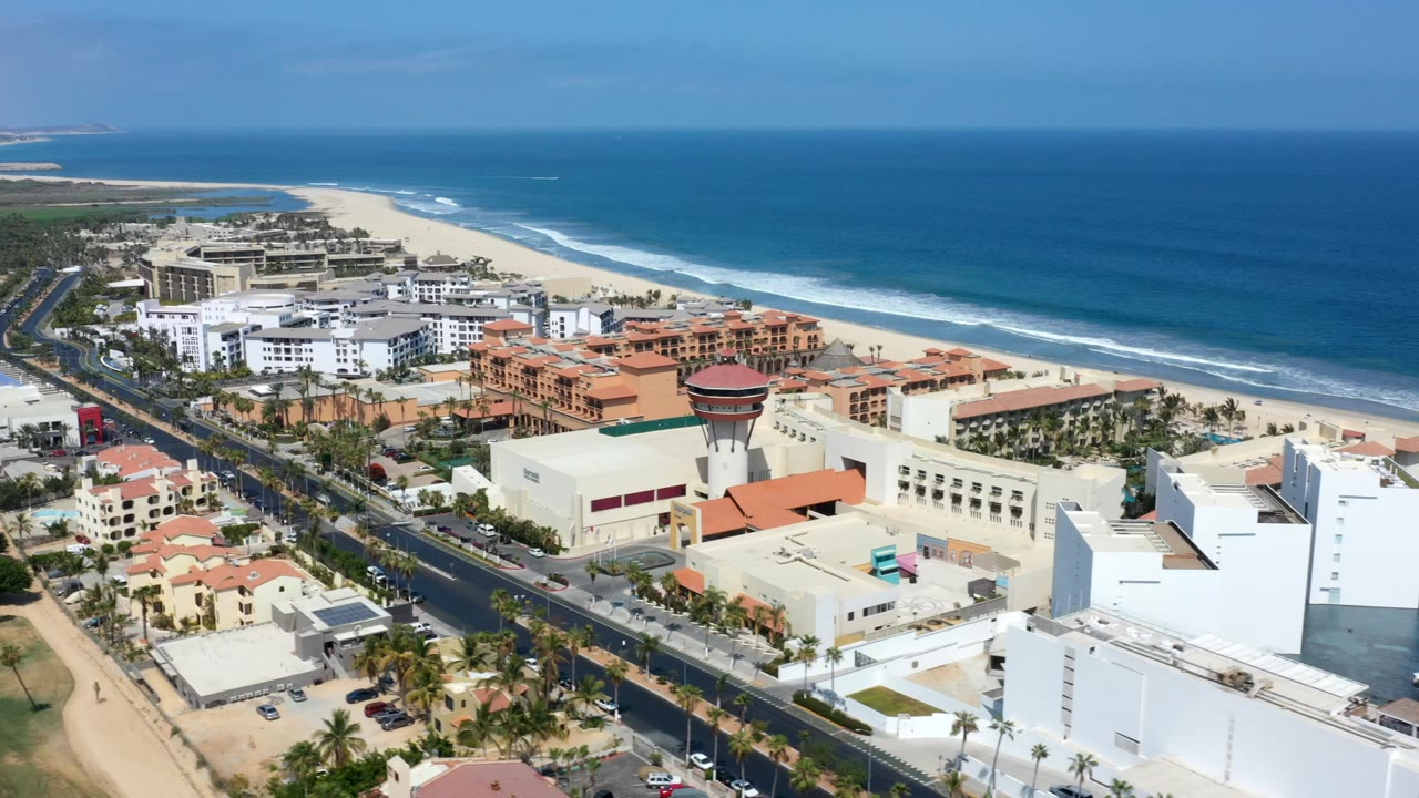 Wide aerial view over Los Cabos, Mexico, showing white and terracotta buildings beside a road, with sandy beach and blue ocean beyond, no visible people.