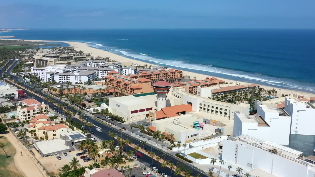 Elevated Los Cabos view with the beachfront road angled through the frame, resort-style buildings below, and white surf bands along the beach, no visible people.