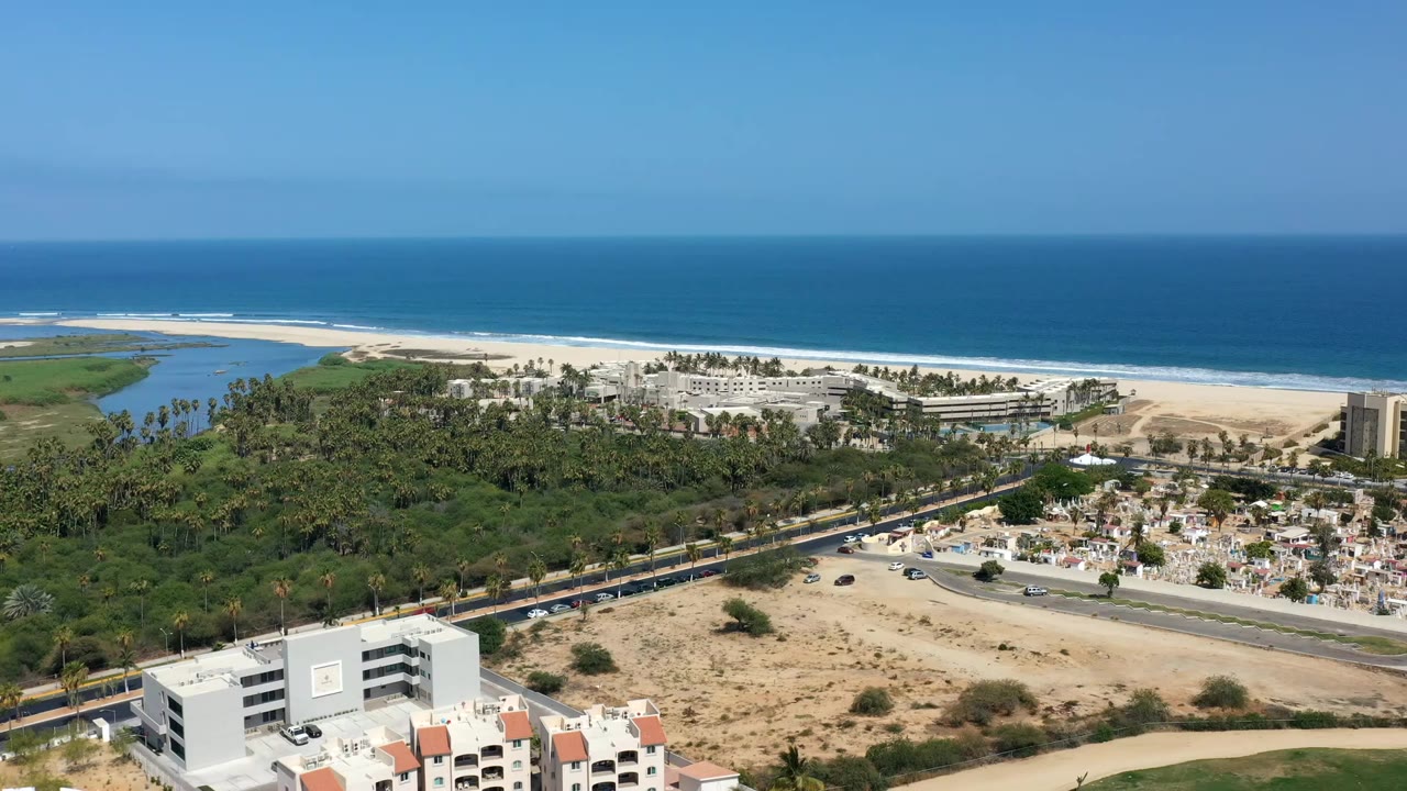Aerial view of ocean, beach, palms, buildings, road, and foreground cemetery area.