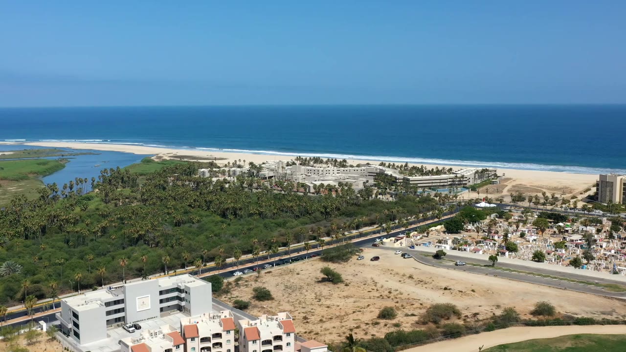 Los Cabos aerial view one second in, showing the lagoon at left, long beach and surf behind low buildings, with the road and sandy foreground area lower in frame.