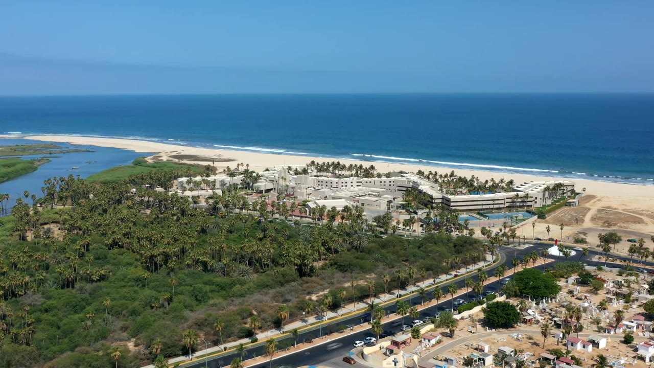 Los Cabos aerial view with the horizon high in frame, surf lines on the beach, palms around low buildings, a curved road in the foreground, vehicles, and no visible people.