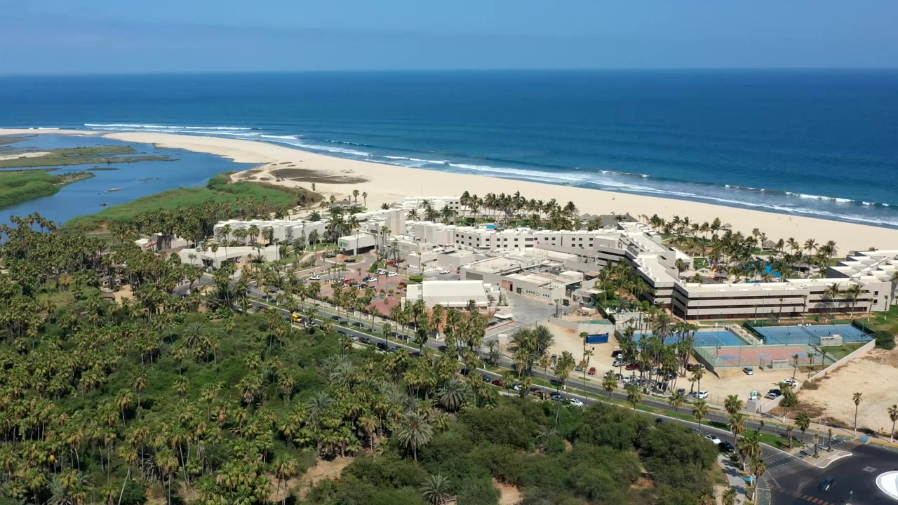 Aerial view in Los Cabos with white resort buildings centered below, tennis courts and roads in the foreground, and a sandy beach, inlet, and ocean behind them.