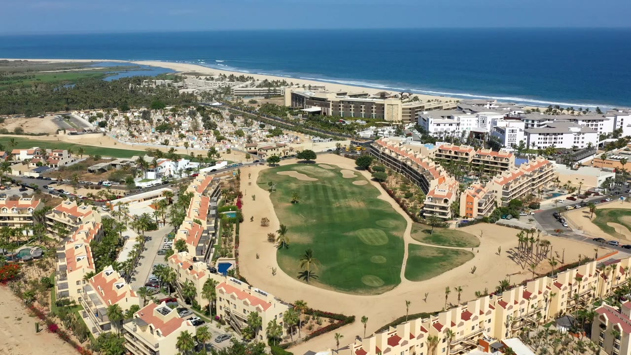Los Cabos aerial view with the golf course centered lower in frame, apartment-style buildings on both sides, sandy beach beyond, and no visible people.