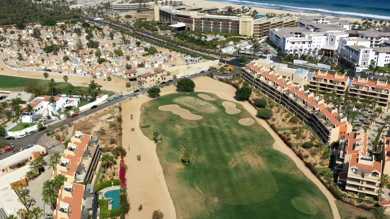 Los Cabos aerial view centered on a green fairway and sand bunkers, with terracotta-roofed buildings on both sides, a road network above, distant surf, and no visible people.