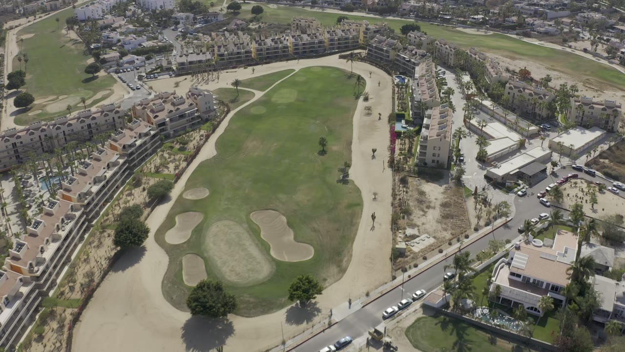 High aerial view of a green course, sand bunkers, roads, buildings, and parked cars.
