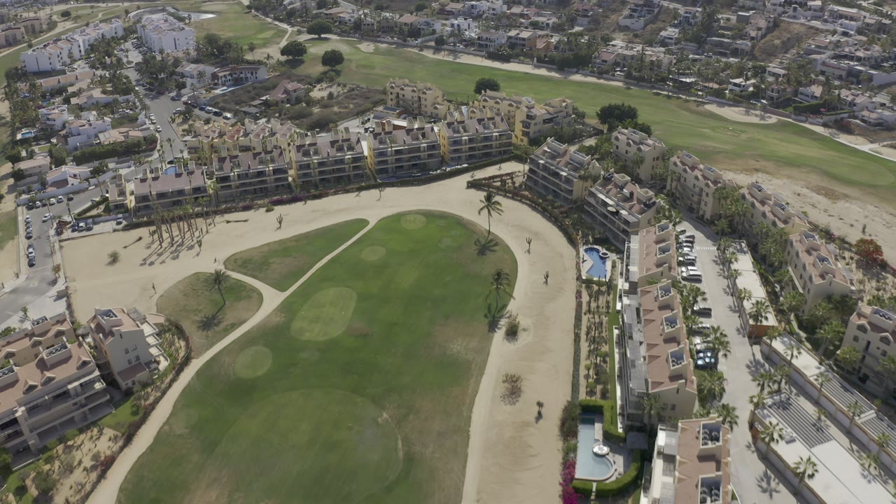 High aerial view in Los Cabos, Mexico, showing a green golf course, sand bunkers, modern buildings, roads, parked cars, and no visible people at the start of the clip.