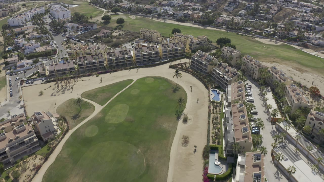 Aerial frame over Los Cabos, Mexico, with the golf green and pale bunkers filling the lower frame, rows of modern buildings above, and no visible people.