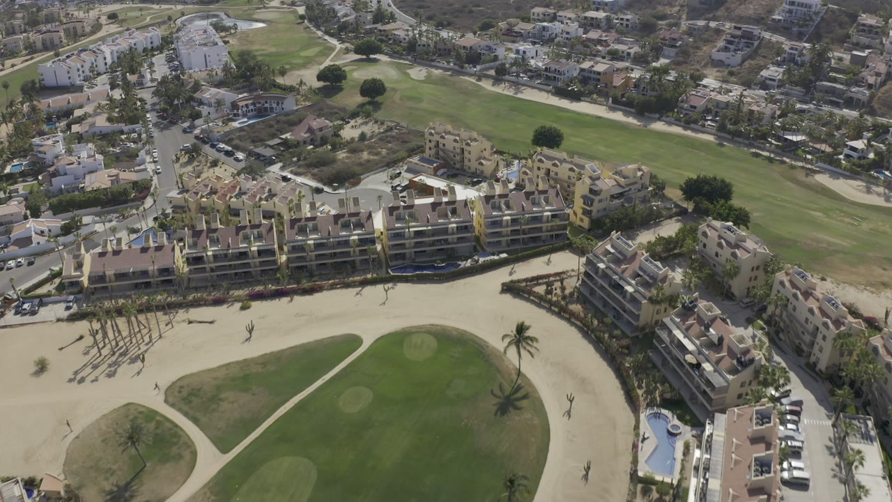 High aerial view in Los Cabos, Mexico showing pale condo buildings, green maintained grounds, sand paths, surrounding roads, and no visible people.