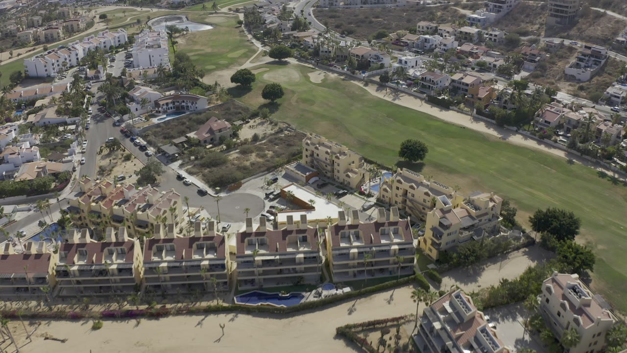 High aerial view over Los Cabos residential buildings, a curving road, parked cars, and a green golf fairway with no visible people.