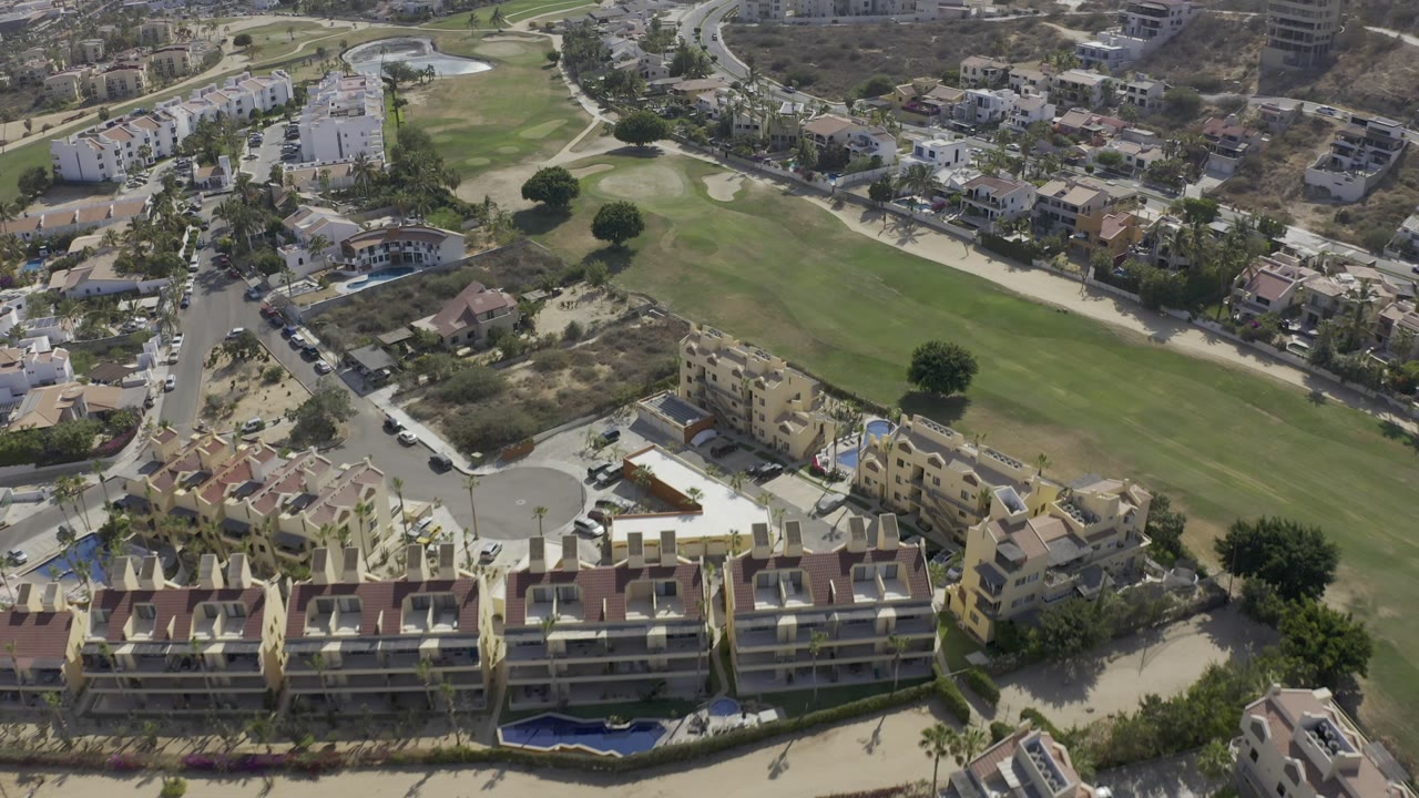 Los Cabos aerial view with villas and apartment blocks around a curved street, green fairway, sand bunkers, and dry hillside lots with no visible people.