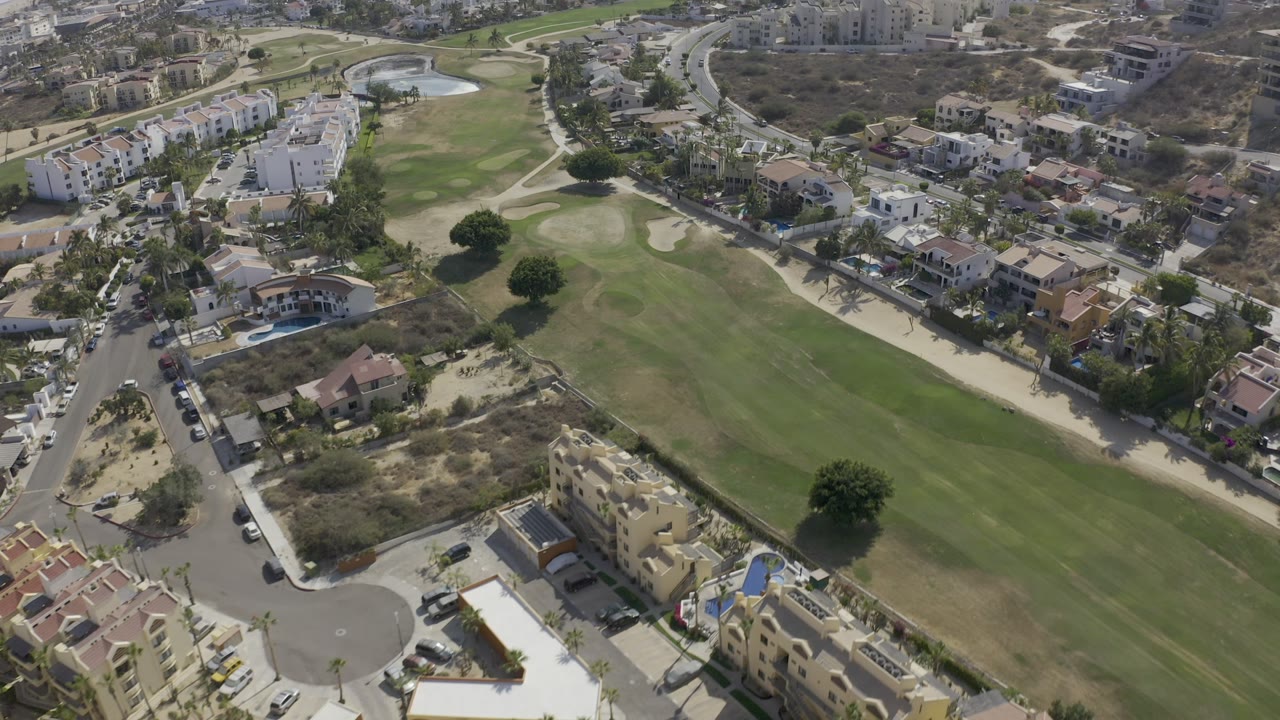Aerial Los Cabos frame showing the fairway centered diagonally, with villas, apartment blocks, roads, and no visible people.