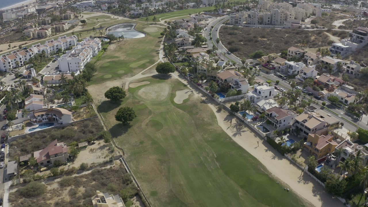 Elevated Los Cabos view with the golf course running through the left side, a curving road to the right, compact homes, and no visible people.