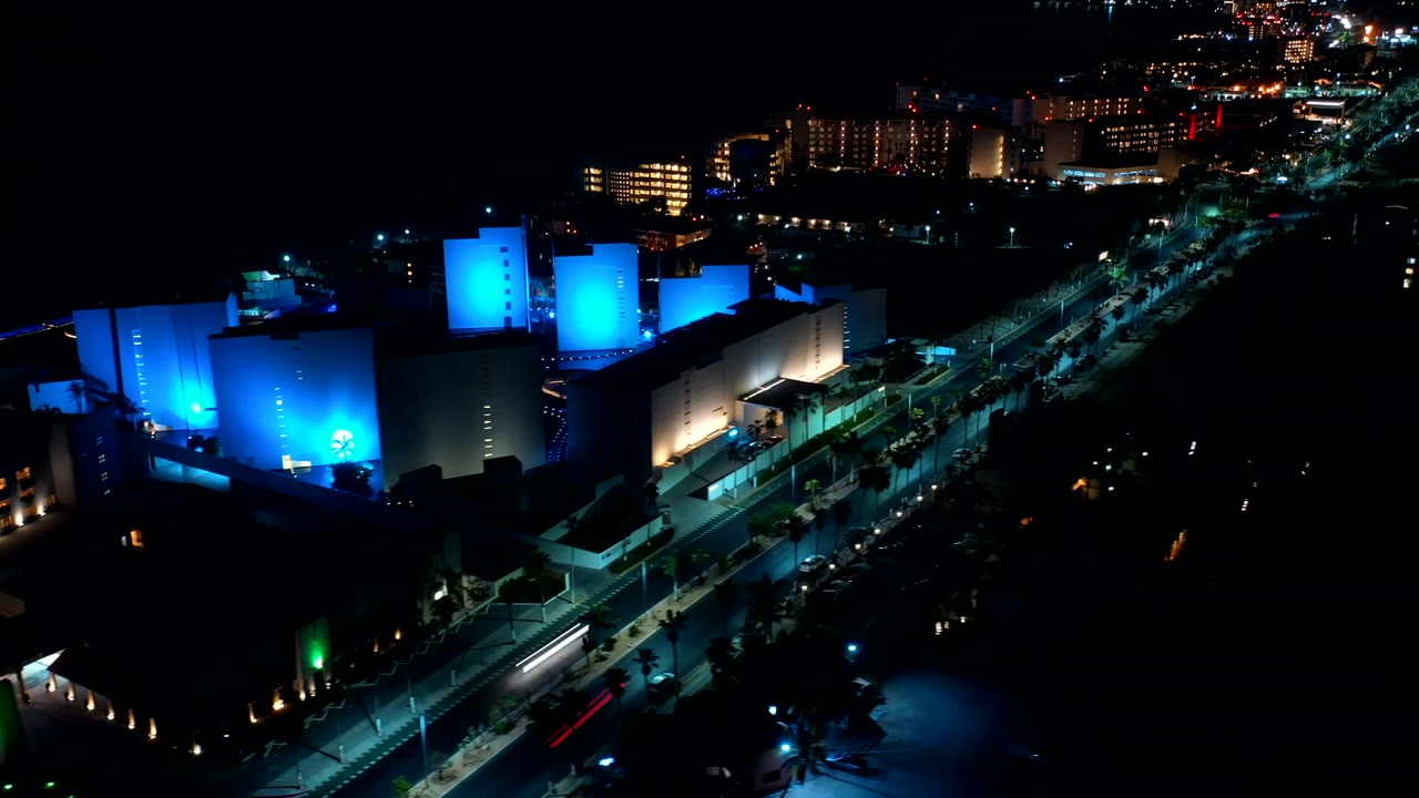 At 0:01, the aerial view holds on the illuminated building faces, with a diagonal road below and no visible people in the dark street areas.