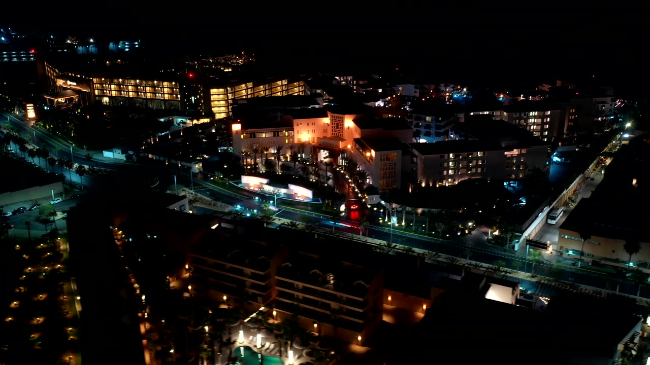 Drone-height night view in Los Cabos, Mexico showing a bright building cluster, dark foreground rooftops, and small vehicle light traces with no visible people.