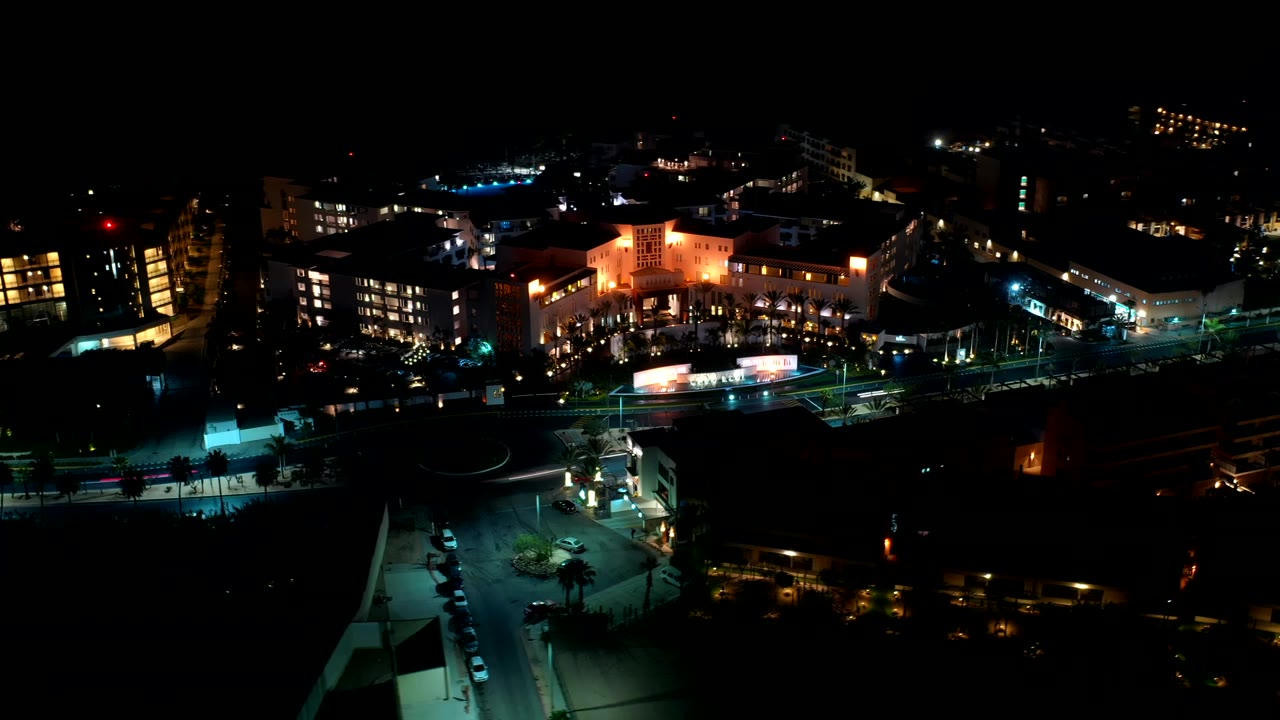 Los Cabos nighttime aerial frame with bright building windows, a lit roadway across the lower frame, and no visible people.